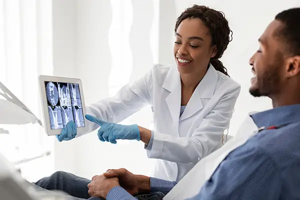 A dentist showing and explaining a dental X-ray image to a smiling male patient on a digital tablet during a consultation.
